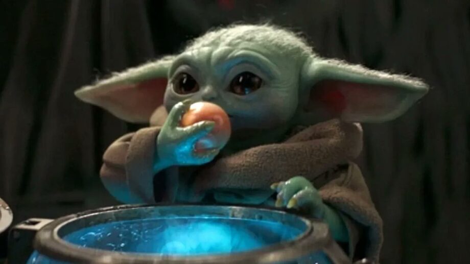 Grogu, pequeno alienígena verde com orelhas grandes e olhos escuros, aparece usando um manto marrom enquanto come um tipo de ovo redondo e alaranjado com a mão. Ele está diante de um recipiente metálico que emite uma luz azul, olhando para frente enquanto leva a comida à boca. O fundo é escuro e desfocado, sugerindo o interior de uma nave ou ambiente fechado.